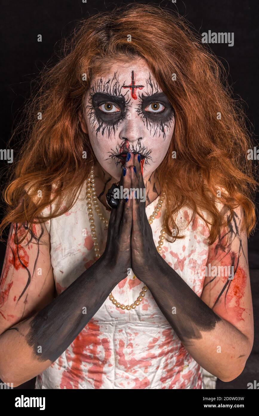 A terrifying redheaded woman with red and white makeup to look creepy with her hands in prayer. Concept Halloween Stock Photo
