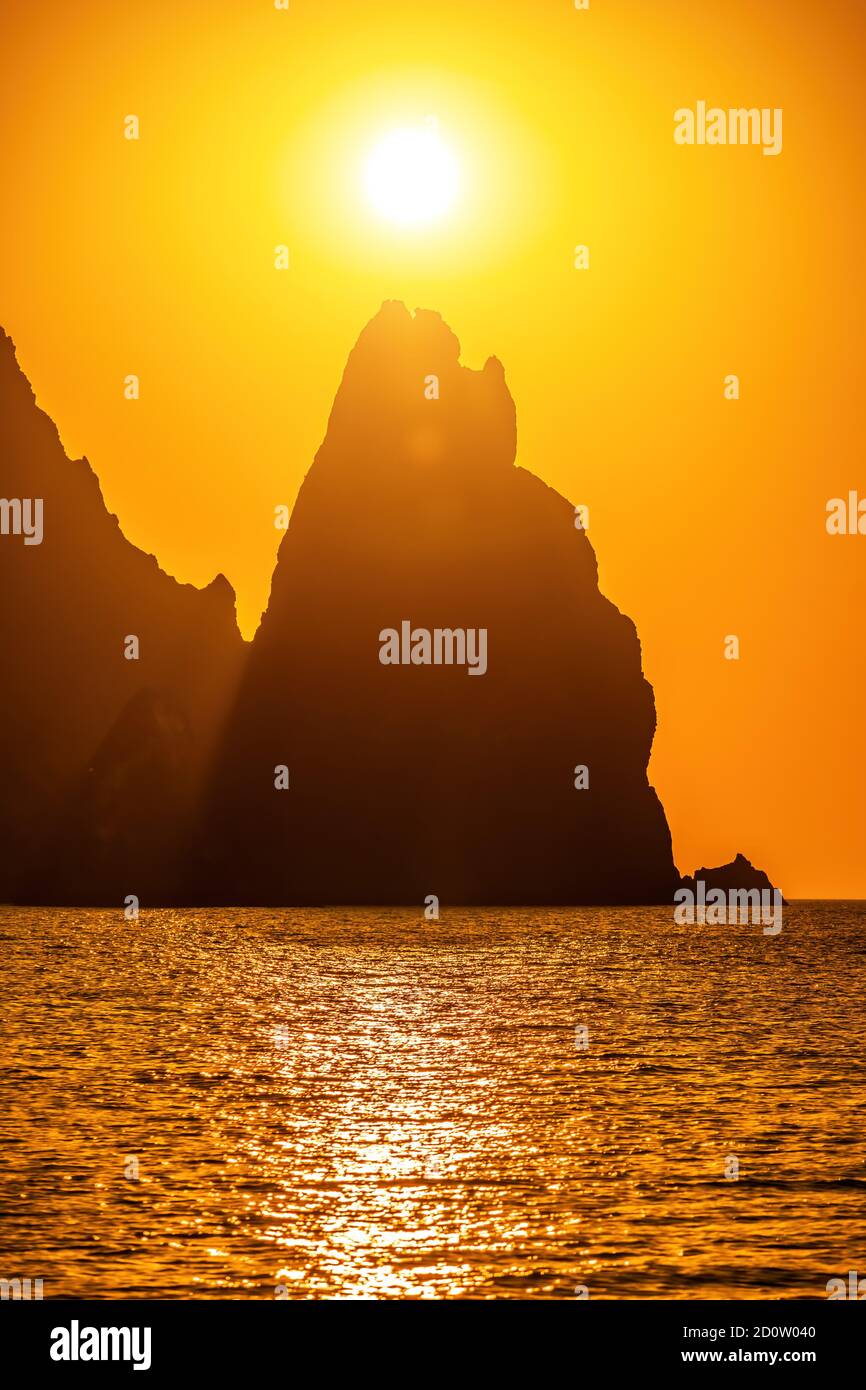 A gold burning sunset with the silhouette of a rocky cape over the sea ...