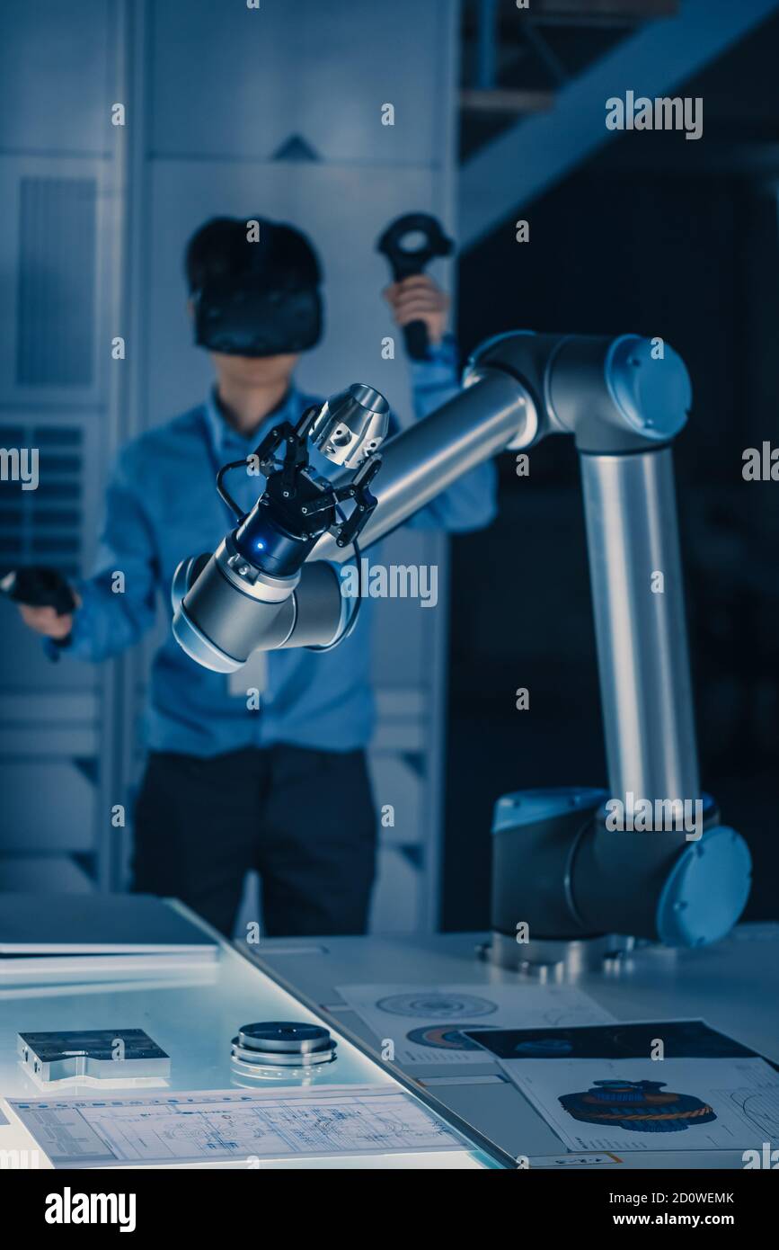 Vertical Shot of Professional Development Engineer in Blue Shirt Controlling Futuristic Robotic Arm with Virtual Reality Headset and Joysticks in a Stock Photo