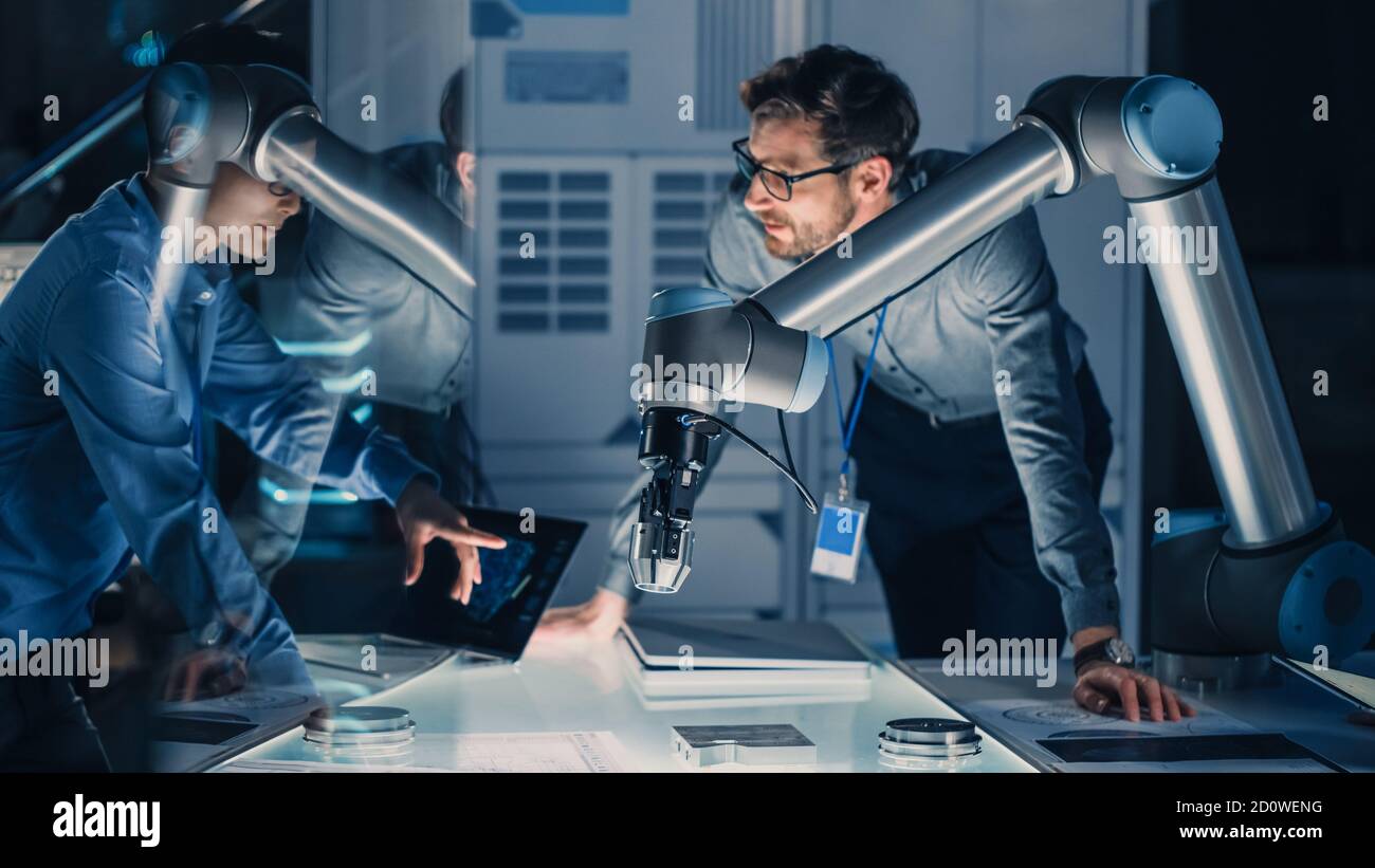 Diverse Team of Engineers with Laptop and a Tablet Analyse and Discuss How a Futuristic Robotic Arm Works and Moves a Metal Object. They are in a High Stock Photo