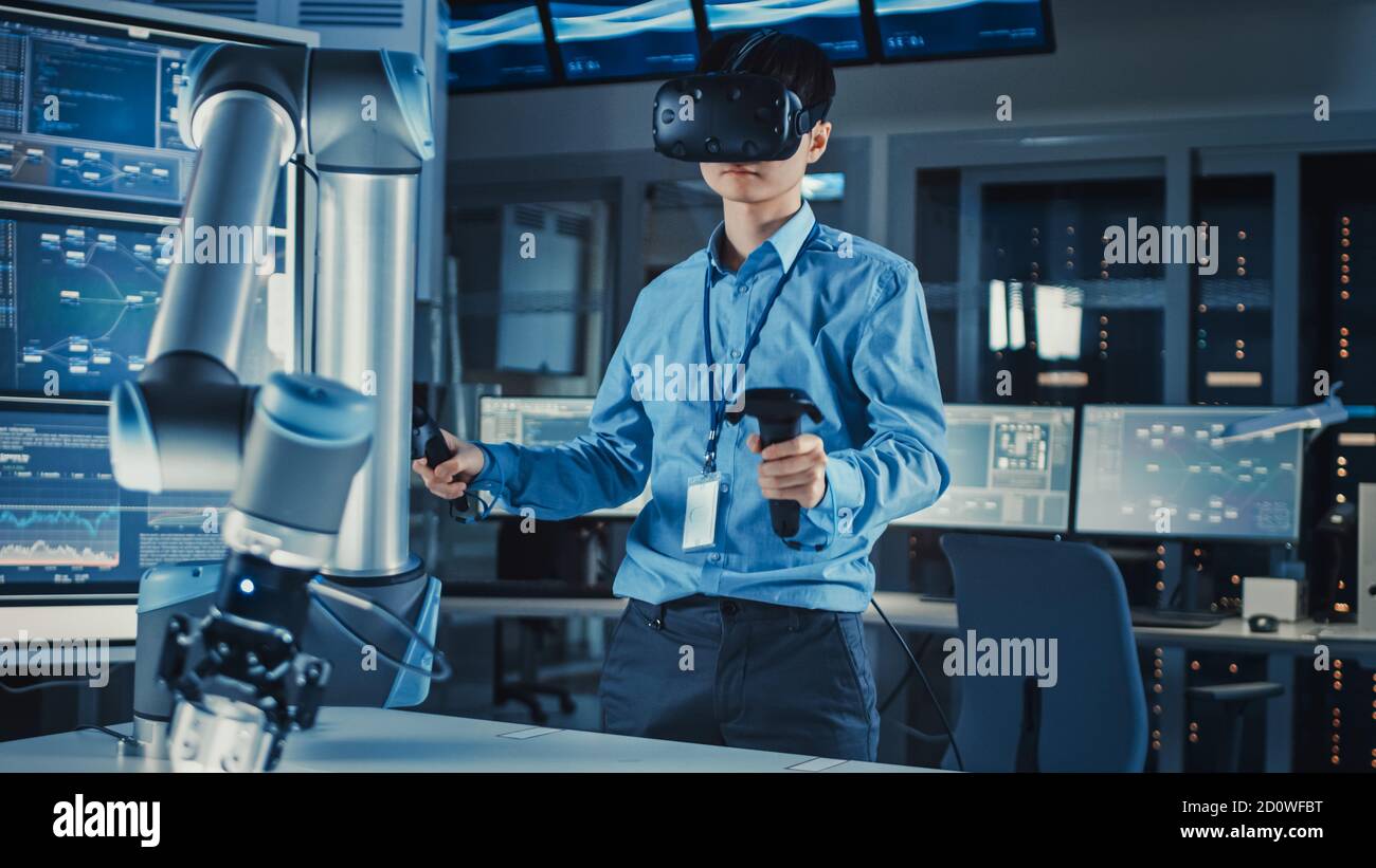 Professional Japanese Development Engineer in Blue Shirt is Controlling a Futuristic Robotic Arm with a Virtual Reality Headset and Joysticks in a Stock Photo