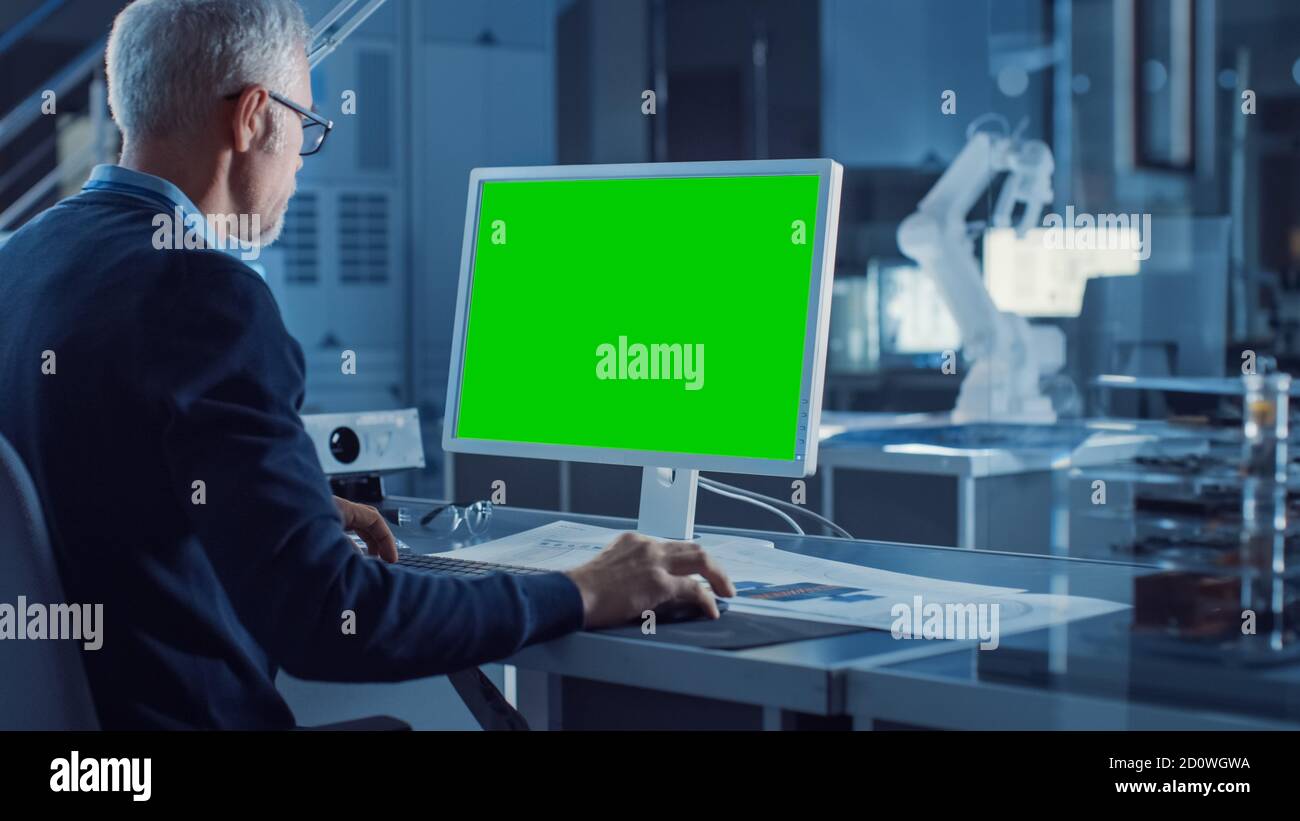 Professional Heavy Industry Engineer Works on Green Mock-up Screen Computer. Engineering Bureau and Industrial Design Agency with Various Robotic Stock Photo