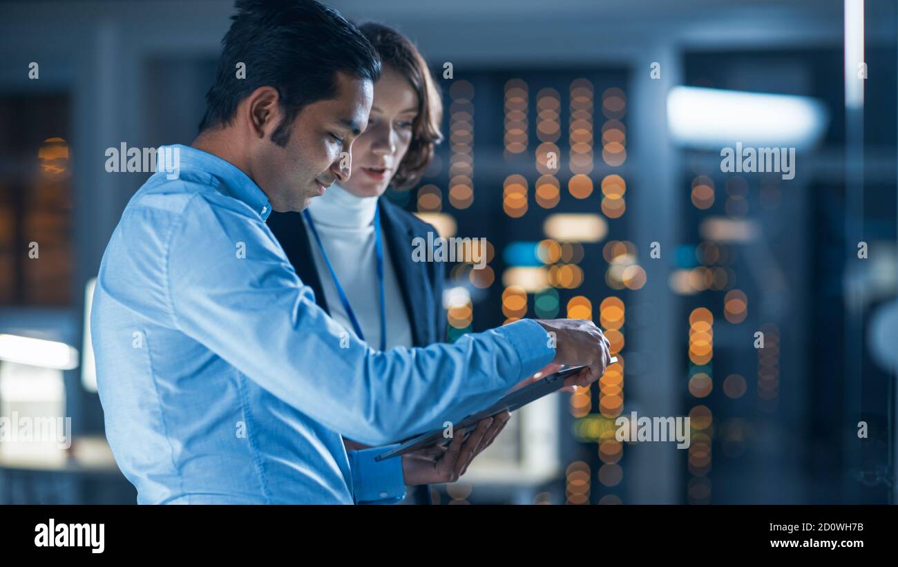 In Technology Research Facility: Female Project Manager Talks With Chief Engineer, they Consult Tablet Computer. Team of Industrial Engineers Stock Photo