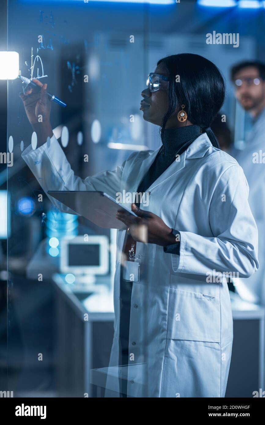 In the Research Laboratory Smart and Beautiful African American Female Scientist Wearing White Coat and Protective Glasses Writes Formula on Glass Stock Photo