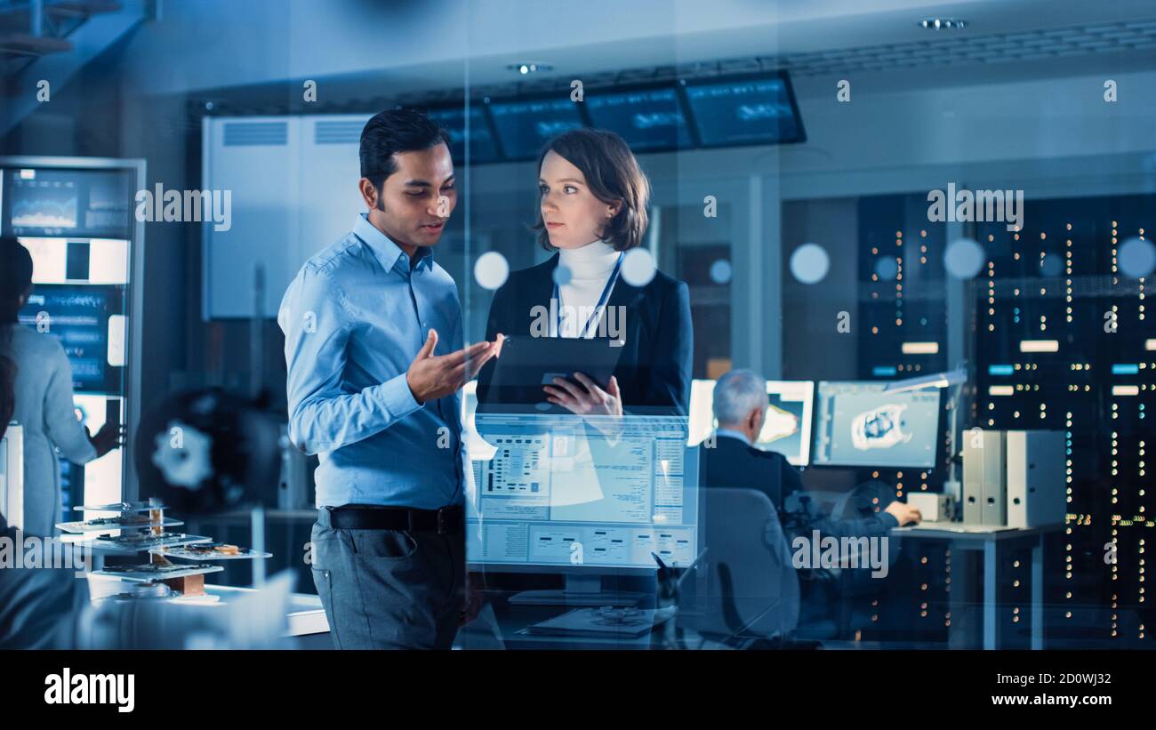 In Technology Research Facility: Female Project Manager Talks With Chief Engineer, they Consult Tablet Computer. Team of Industrial Engineers Stock Photo