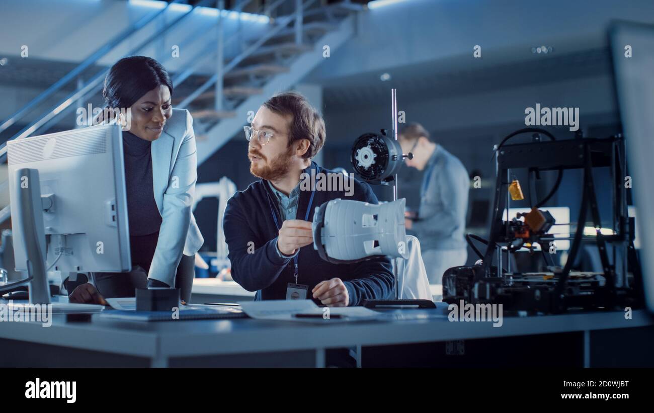 Electronics Development Engineer Working at His Desk, Talks with Project Manager, Shows High Tech Prototype Component. Team of Professionals Working Stock Photo