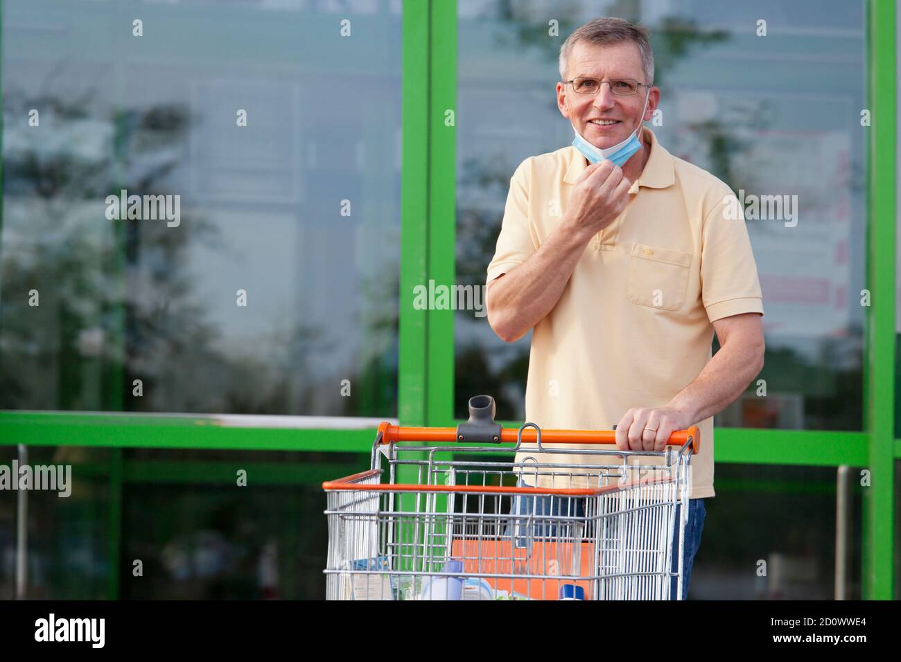 Mature smiling man puts off a face mask while leaving a supermarket with a shopping cart - focus on the face Stock Photo