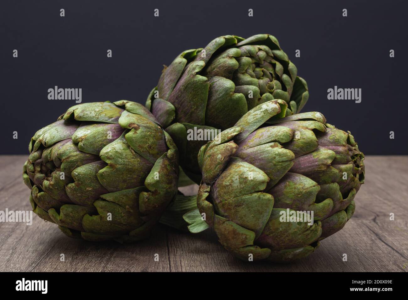 Three fresh organic purple artichokes with stem on a rustic wooden surface and dark background Stock Photo