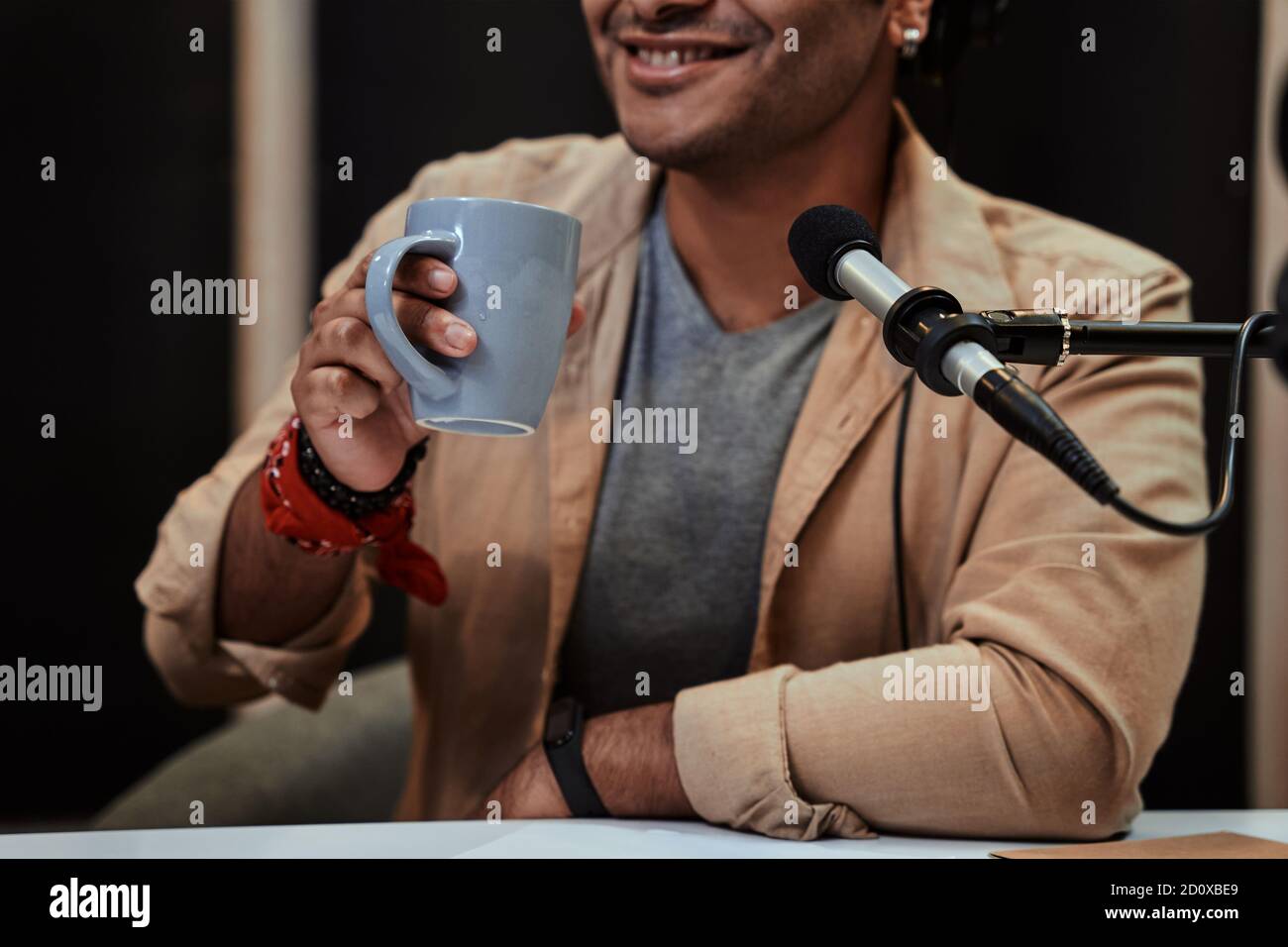 Cropped portrait of young male radio host in headphones smiling, drinking coffee while broadcasting in studio Stock Photo