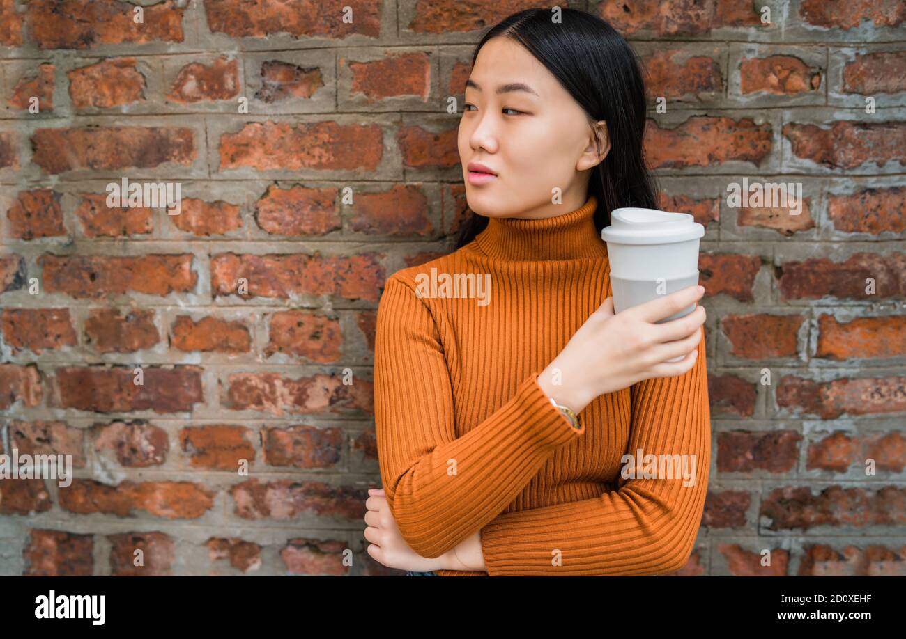 Portrait of young Asian woman holding a cup of coffee against brick ...