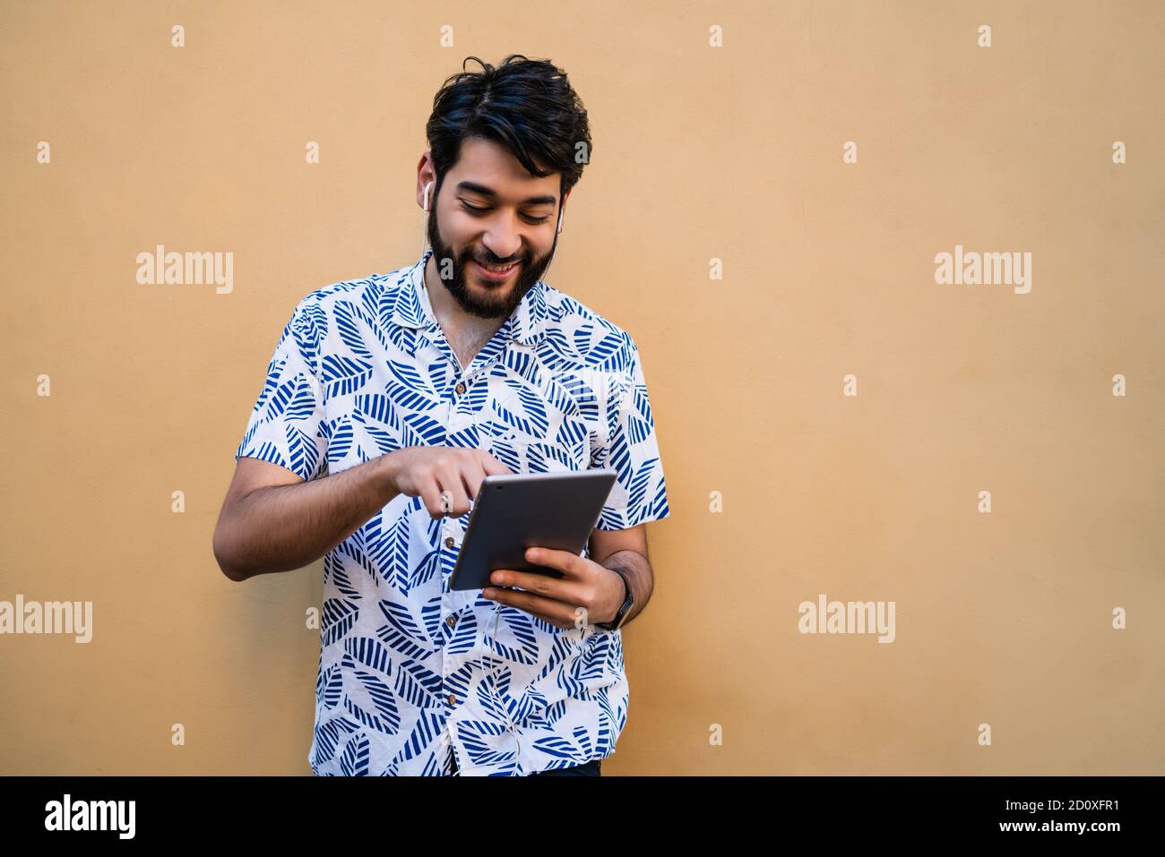 Portrait of young latin man using his digital tablet with earphones against yellow wall. Technology and urban concept. Stock Photo