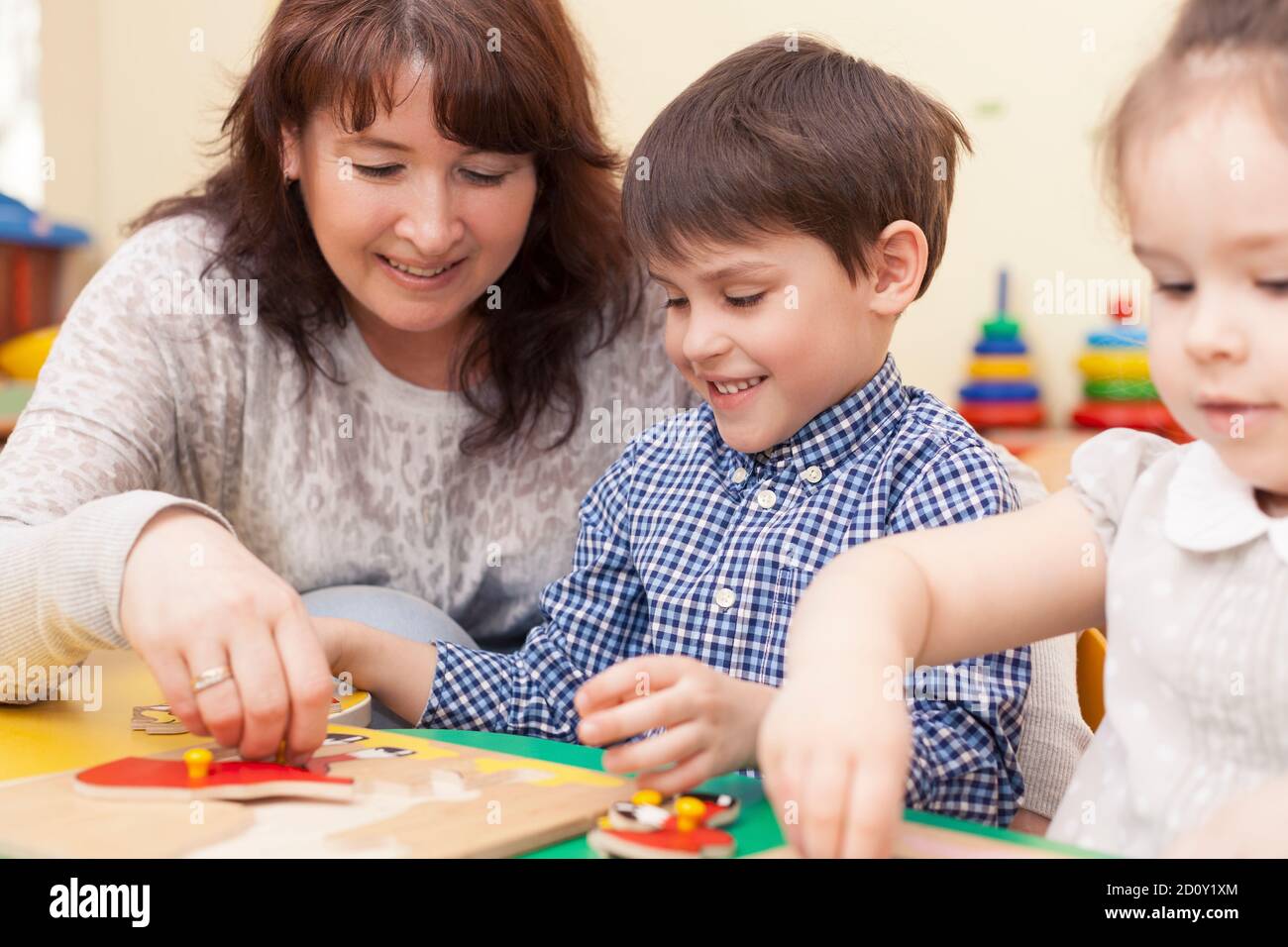 beautiful mature caucasian female teacher collects the puzzle with a pupil,  a boy of preschool age. In the classroom, at the green table. Stock Photo