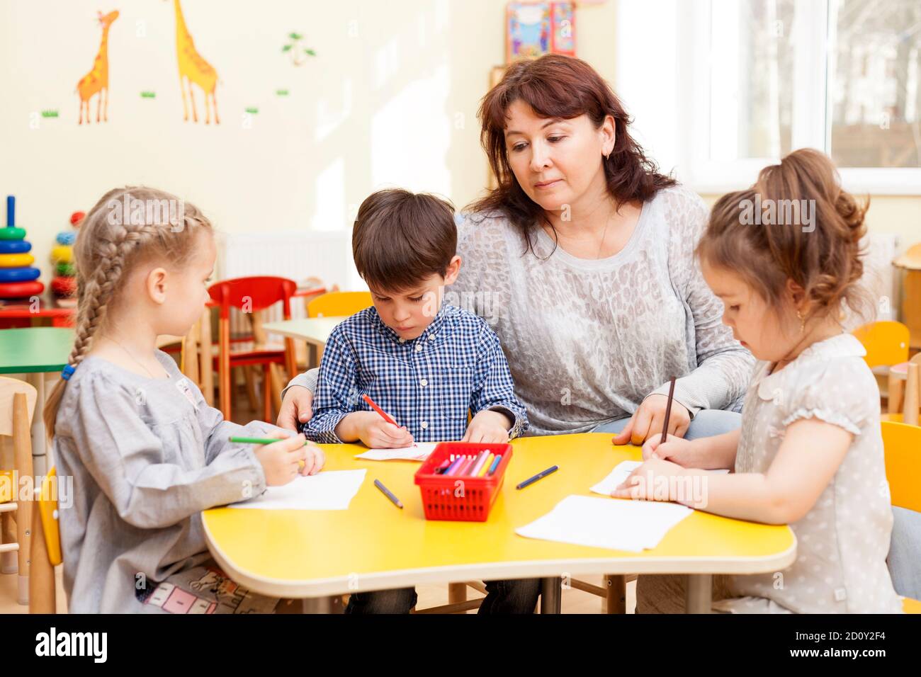 Three preschooler, two girls and a boy, paint pencils at the table. Mature, handsome Caucasian brunette woman, caregiver, watching and smiling. Stock Photo