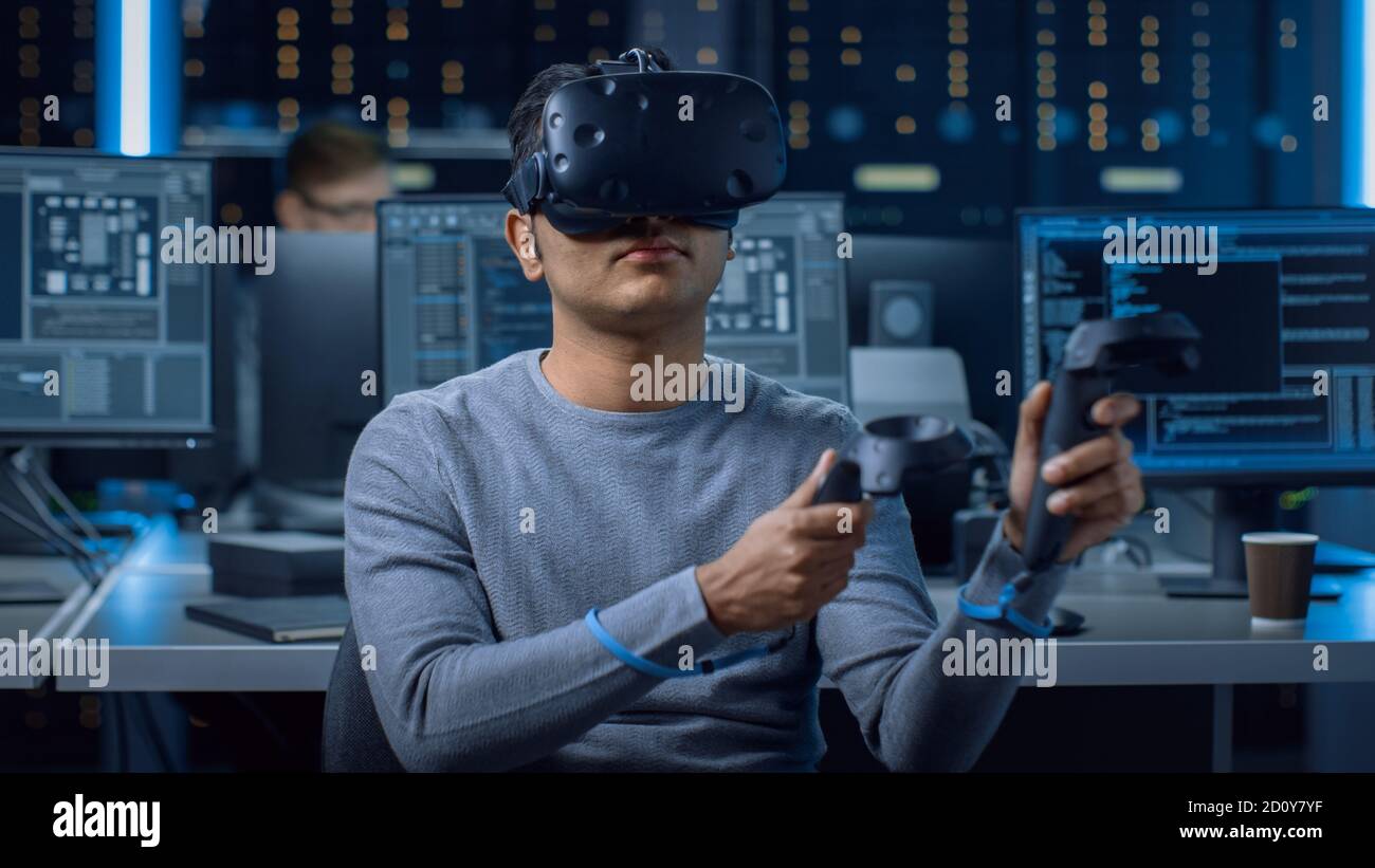 Portrait of Software Delevoper Wearing Virtual Reality Headset Using Controllers to Develop and Program VR Gaming Applications. In Background Stock Photo