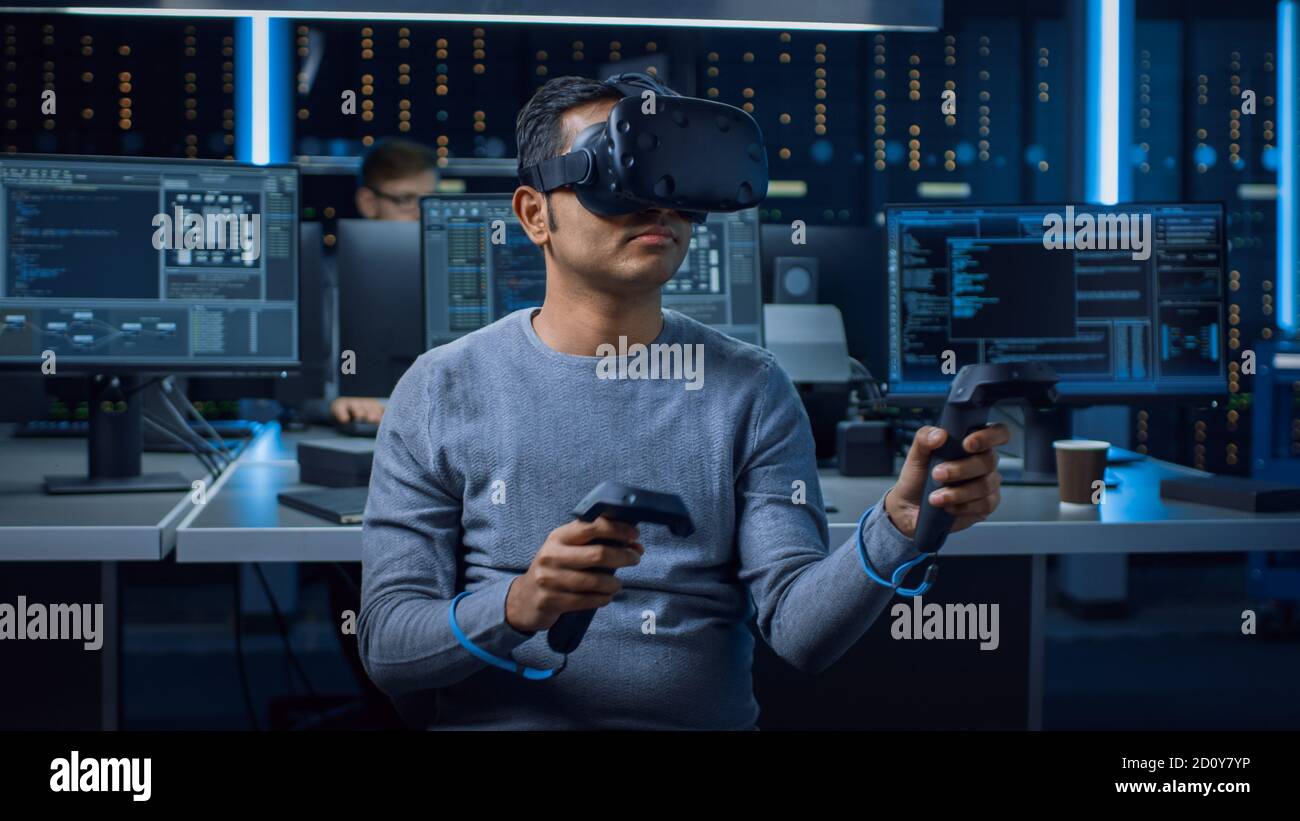 Portrait of Software Delevoper Wearing Virtual Reality Headset Using Controllers to Develop and Program VR Gaming Applications. In Background Stock Photo