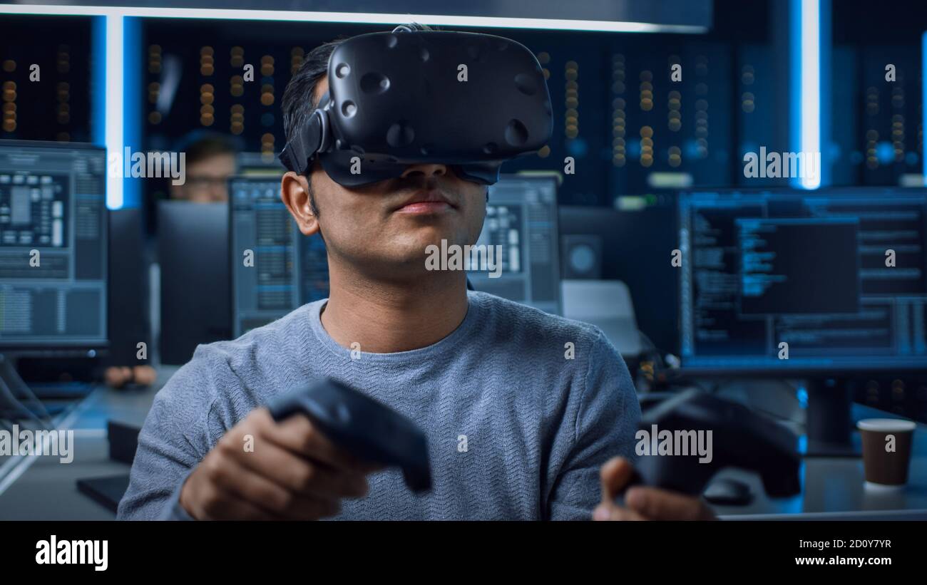 Portrait of Software Delevoper Wearing Virtual Reality Headset Using Controllers to Develop and Program VR Gaming Applications. In Background Stock Photo