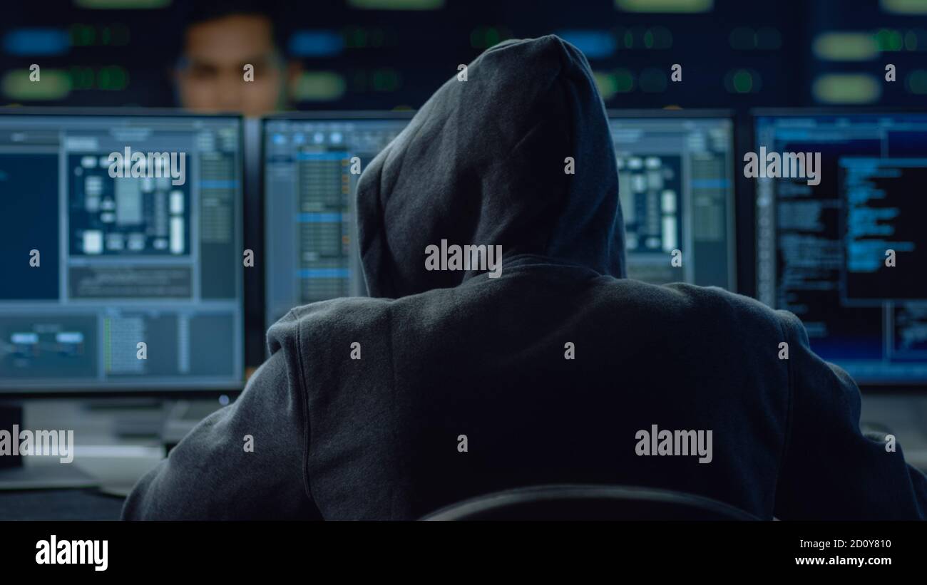Hooded IT Hacker or Specialist Works on a Personal Computer With Display Showing Software Program with Coding Language Interface Stock Photo