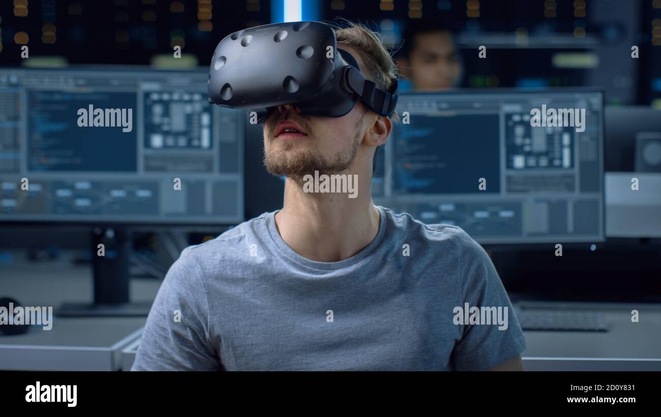 Software Delevoper Wearing Virtual Reality Headset Gesturing with His Head, Developing and Programming VR Game or Application. In Background Stock Photo