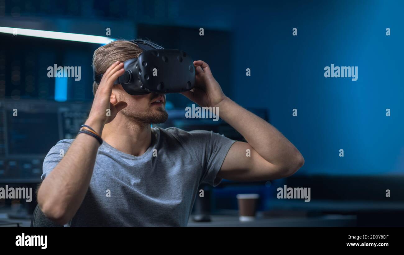 Software Delevoper Putting on Virtual Reality Headset on with His Head, Developing and Programming VR Game or Application. In the Background Stock Photo