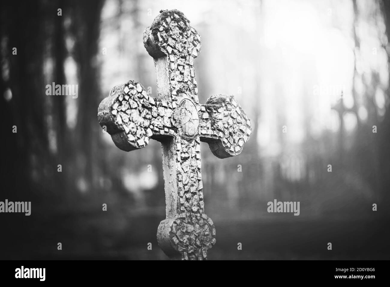 A black-and-white image of an old stone cracked cross standing in the middle of a foggy abandoned scary cemetery. Sadness. Death. Stock Photo