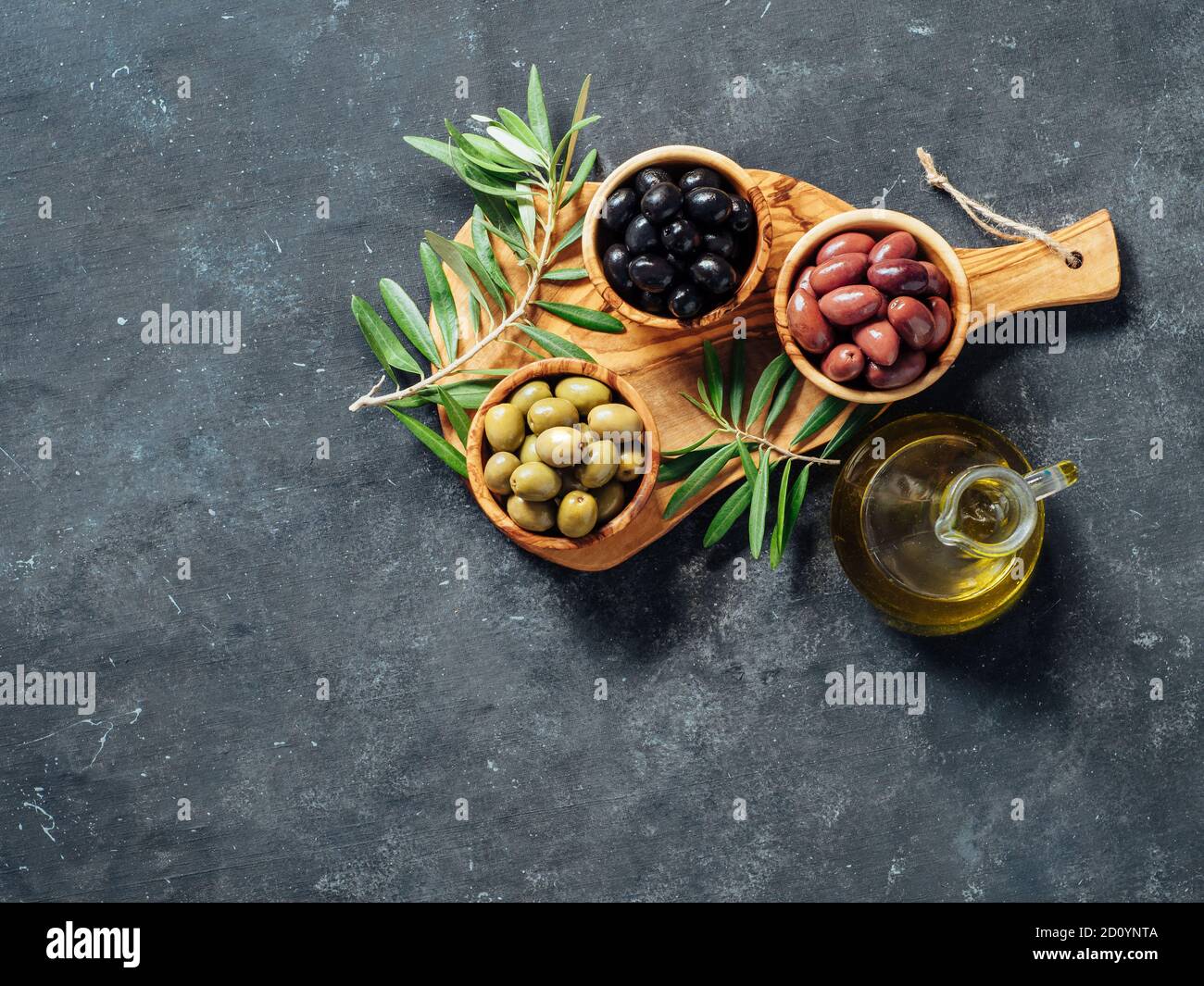 Set of green, black and red or pink olives and olive oil on gray background. Different types of olives in olive wooden bowls and olive oil over wooden cutting board. Copy space. Top view or flat lay Stock Photo