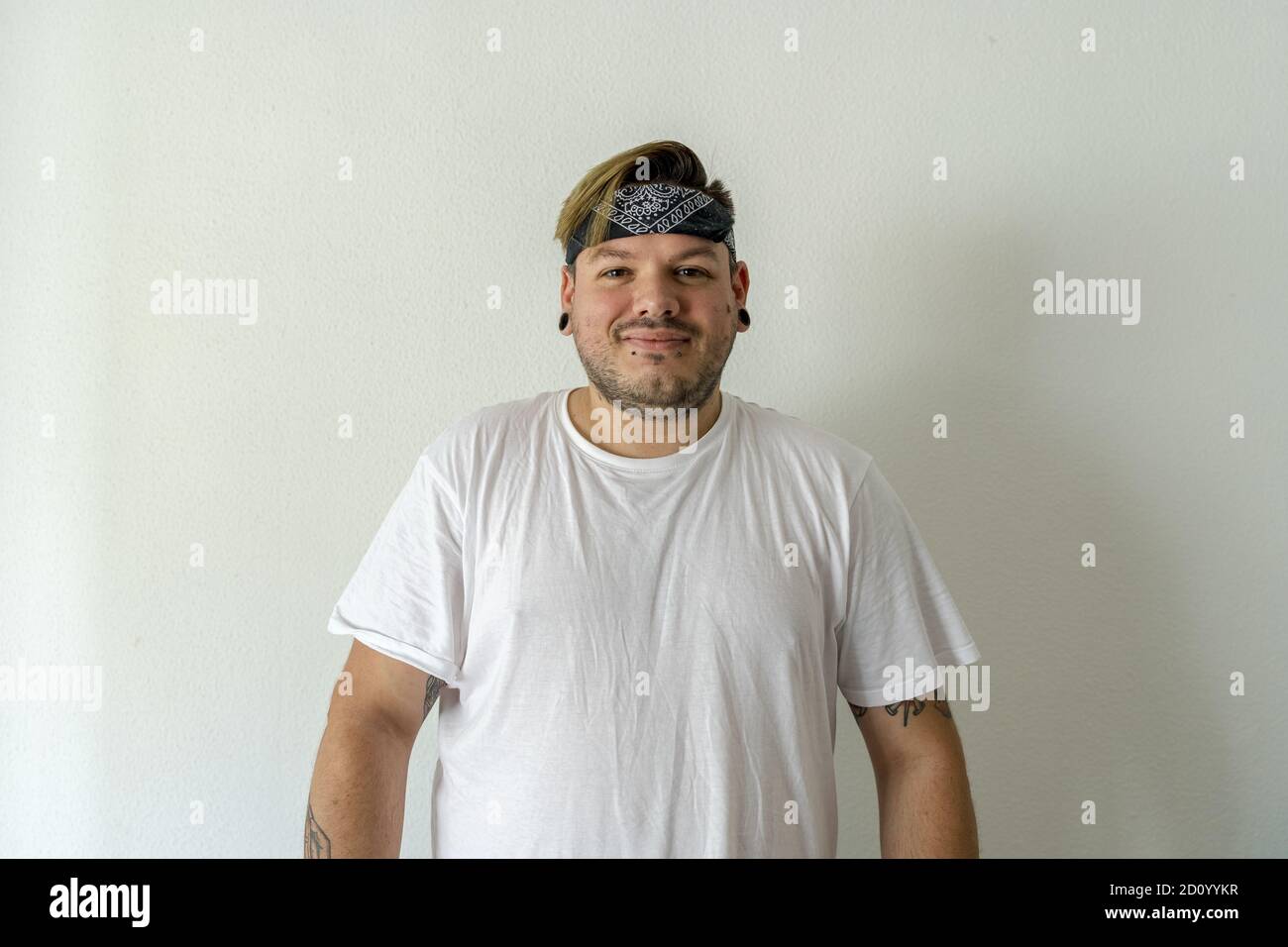 A smiling Caucasian male with tattoos wearing a bandana on his head against a wall Stock Photo