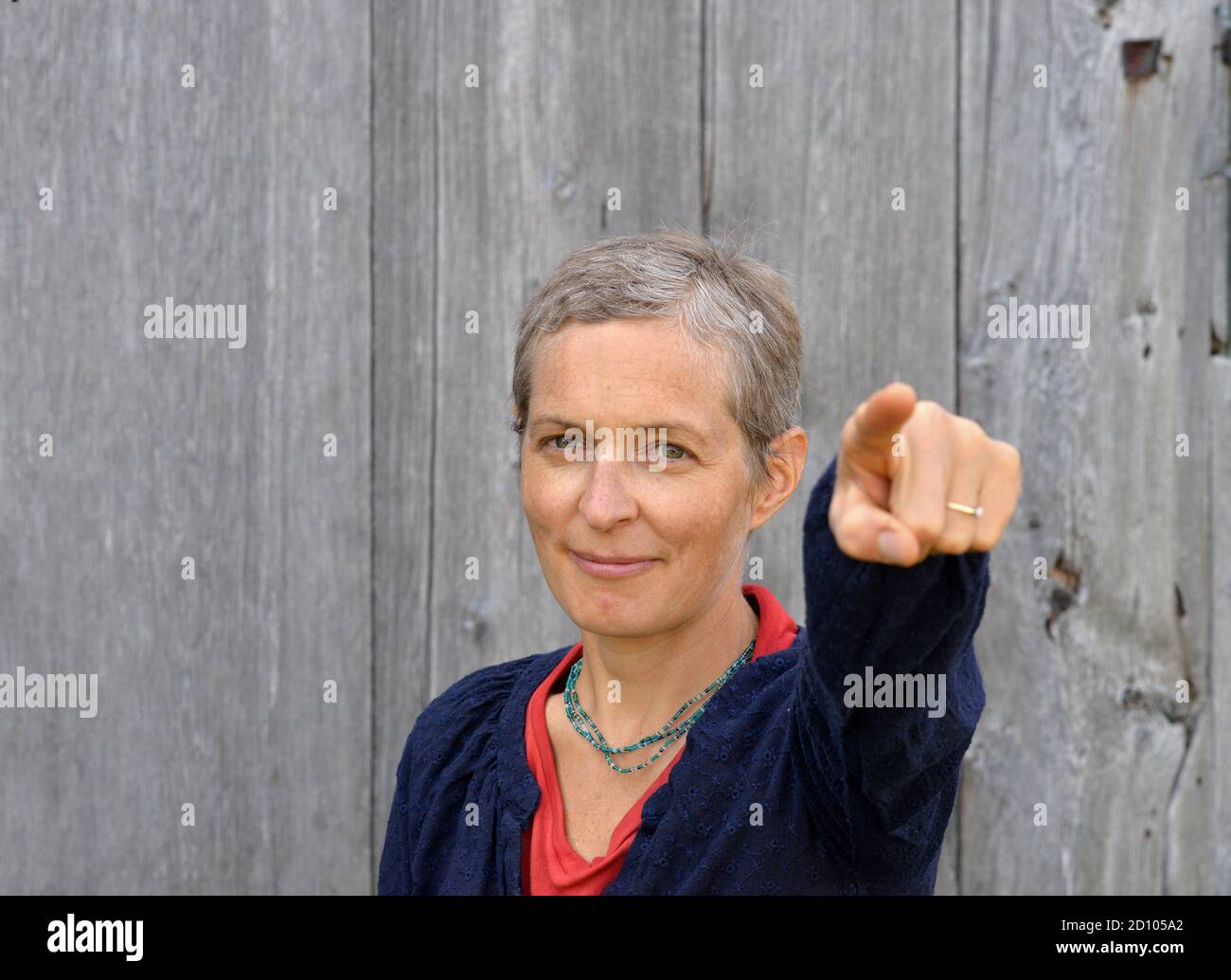 Determined middle-aged Caucasian country woman with short hair points at viewer with index finger of left hand, in front of old barn wood background. Stock Photo