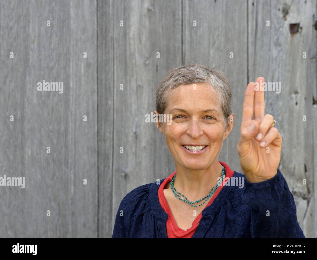 Positive middle-aged Caucasian country woman with short hair (homesteader) takes an oath with two fingers, in front of an old barn wood background. Stock Photo