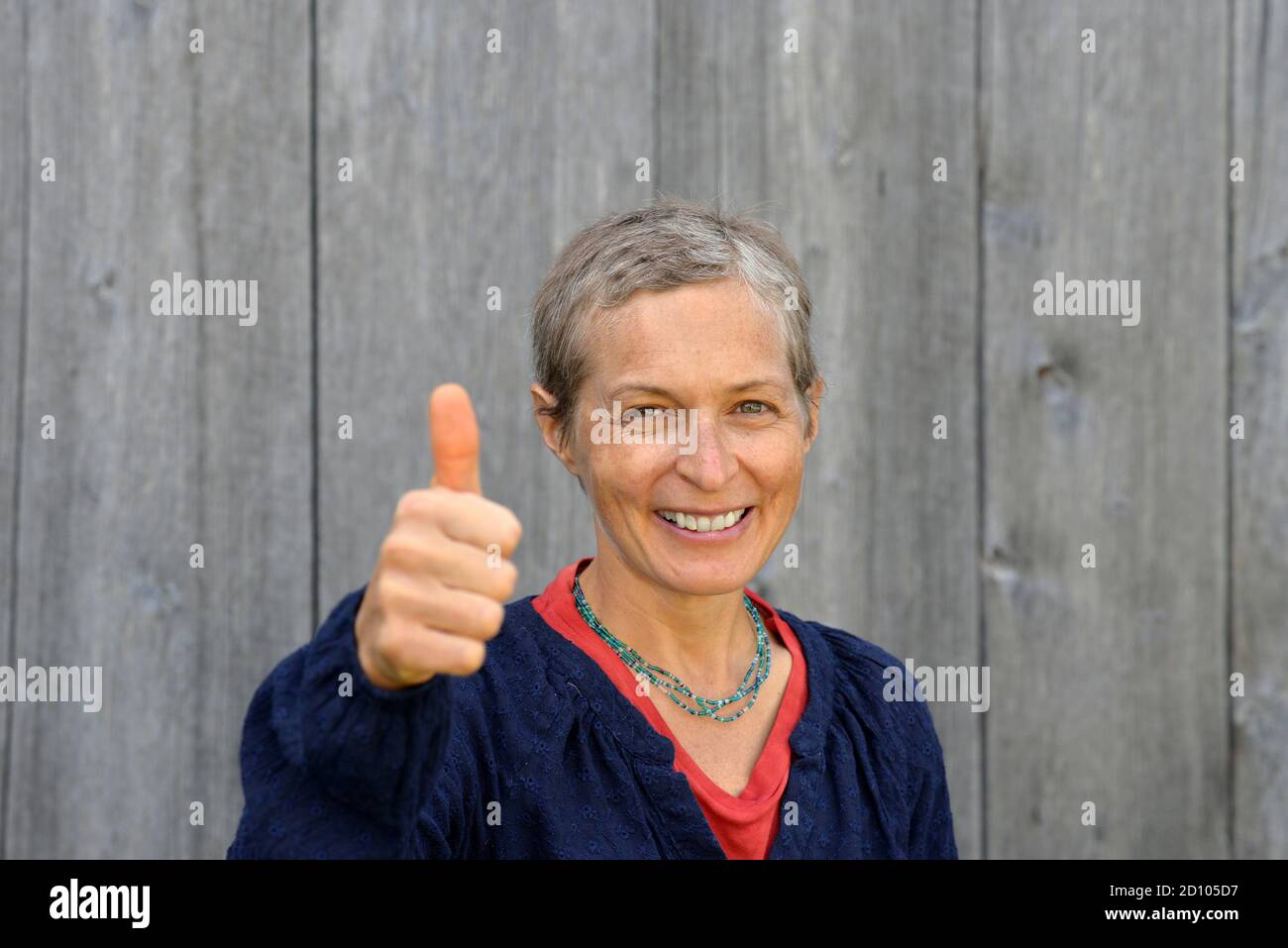 Optimistic middle-aged Caucasian country woman with short hair (modern homesteader) smiles and shows thump up, in front of old barn wood background. Stock Photo