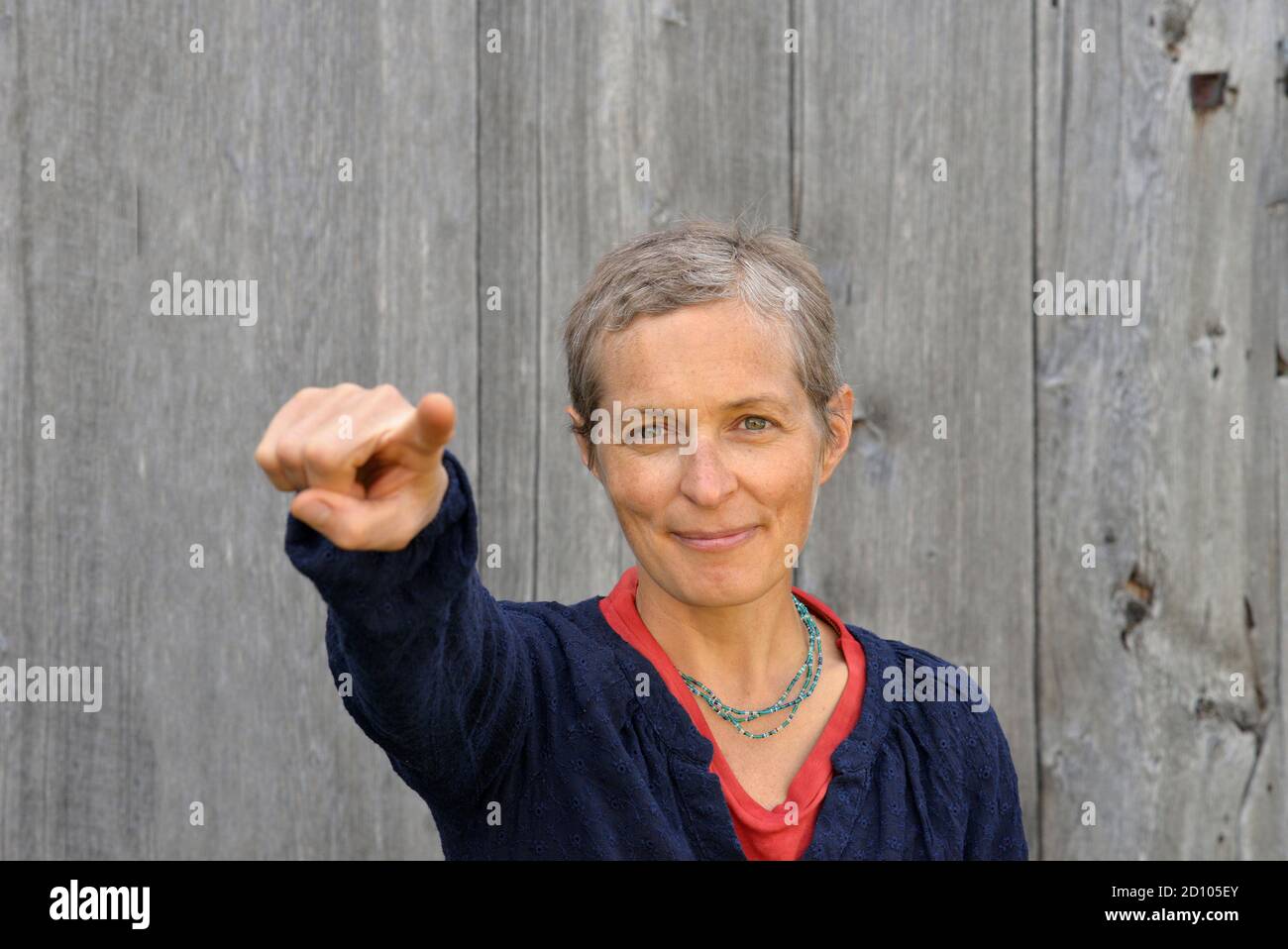 Positive middle-aged Caucasian country woman with short hair points at viewer with index finger of right hand, in front of old barn wood background. Stock Photo