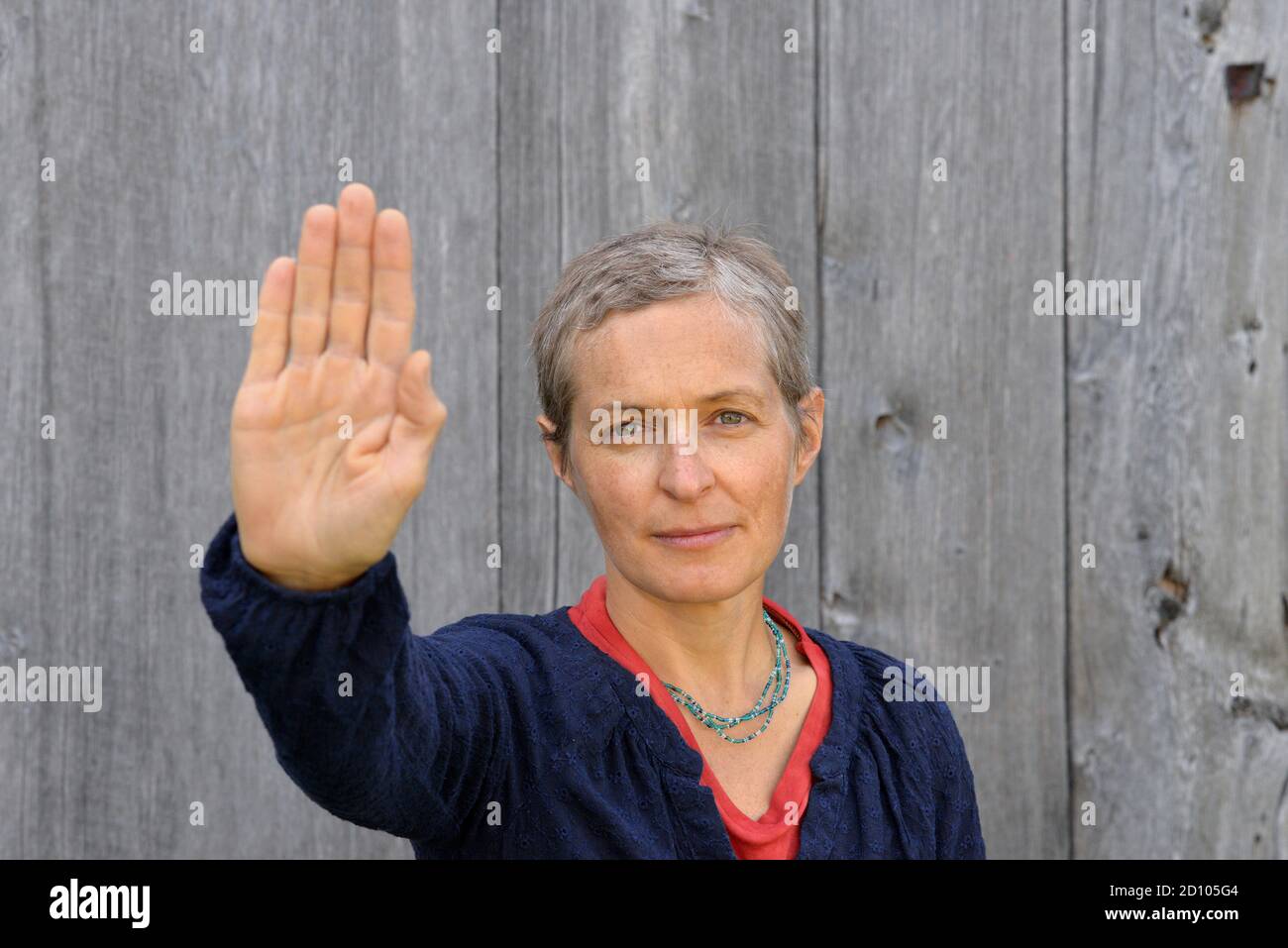 Angry middle-aged Caucasian country woman with short hair makes the hand palm stop sign with her right hand, in front of an old barn wood background. Stock Photo