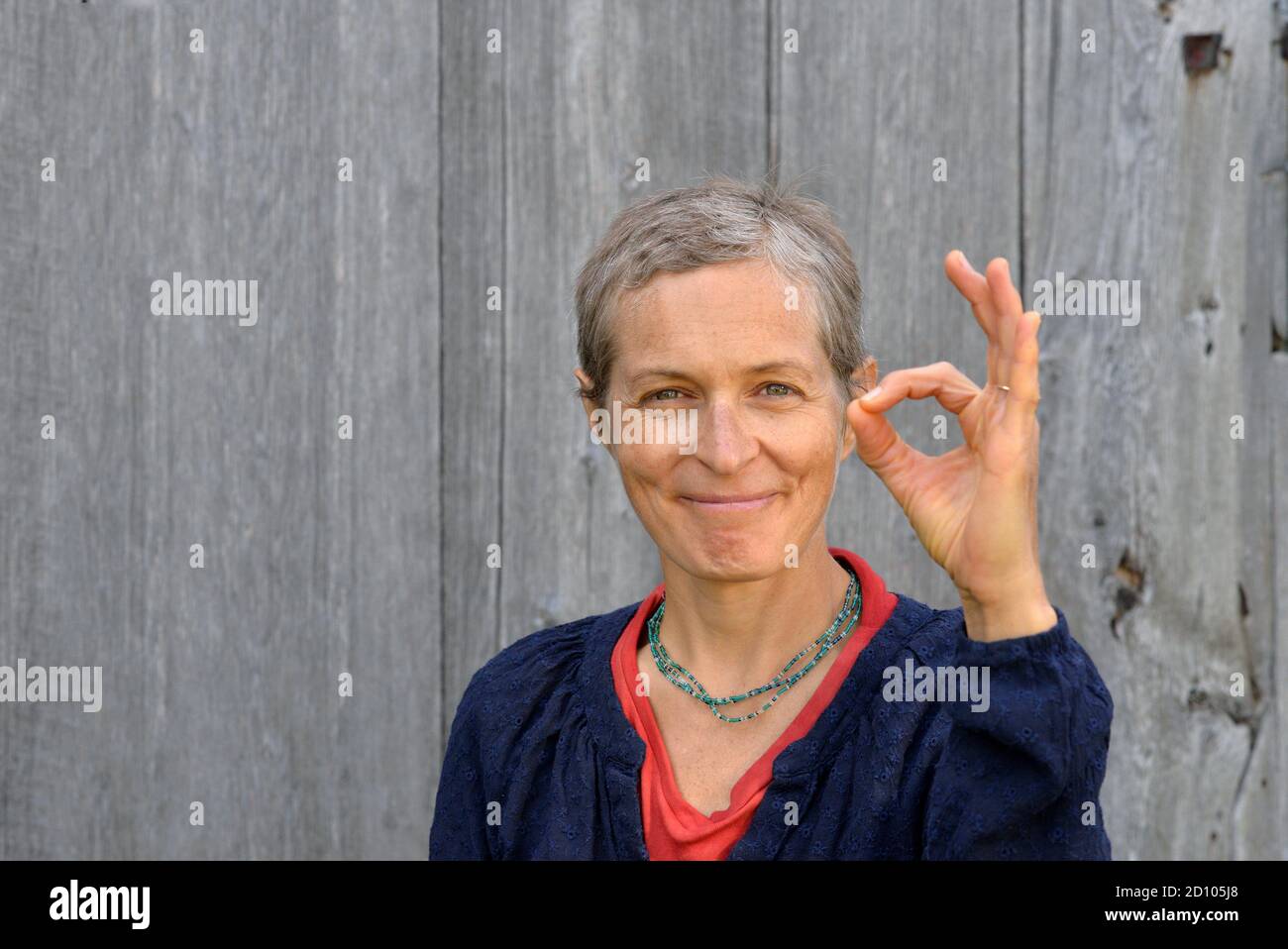 Optimistic modern middle-aged Caucasian country woman shows the ok sign (approval gesture) with her left hand, in front of old barn wood background. Stock Photo