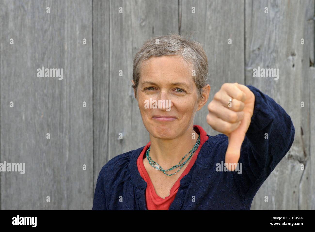 Discontent middle-aged Caucasian country woman with short hair shows thump down (disapproval sign), in front of old barn wood background. Stock Photo