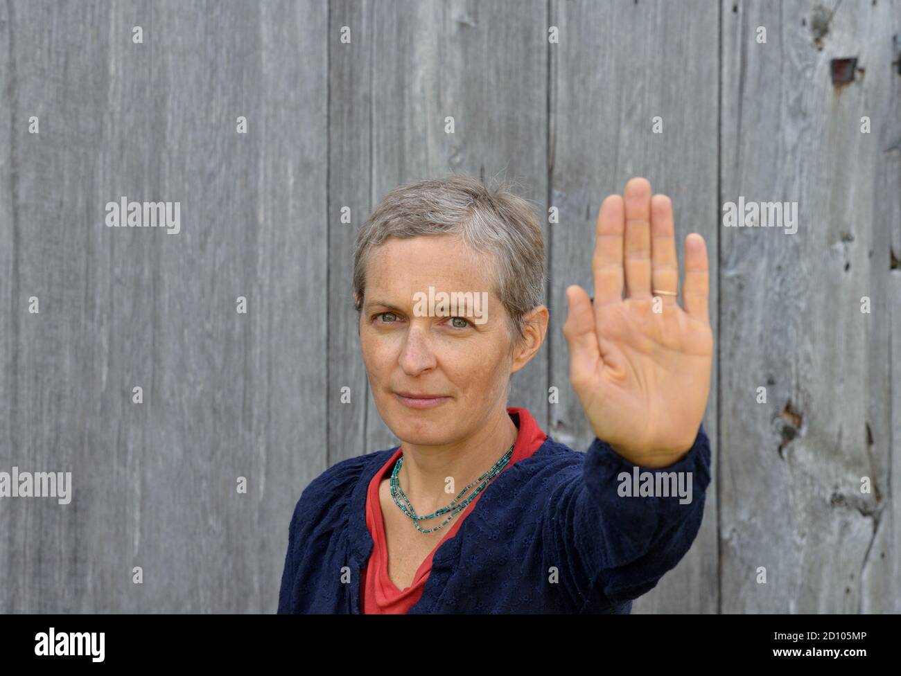 Serious middle-aged Caucasian country woman makes the hand palm stop sign with her left hand (stop gesture), in front of old barn wood background. Stock Photo