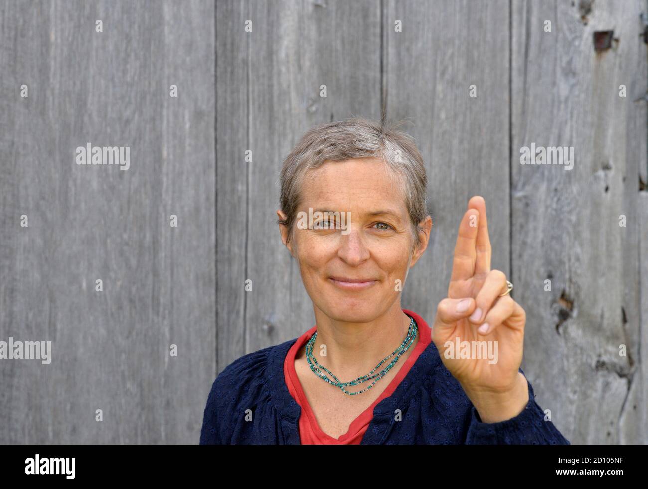 Positive middle-aged Caucasian country woman with short hair keeps her fingers crossed and wishes for good luck, in front of old barn wood background. Stock Photo