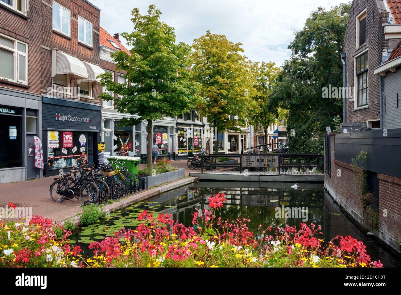 View of the Gouda old city centre. Zeugstraat with canal, various shops and blooming flowers on a sunny afternoon. South Holland, The Netherlands. Stock Photo
