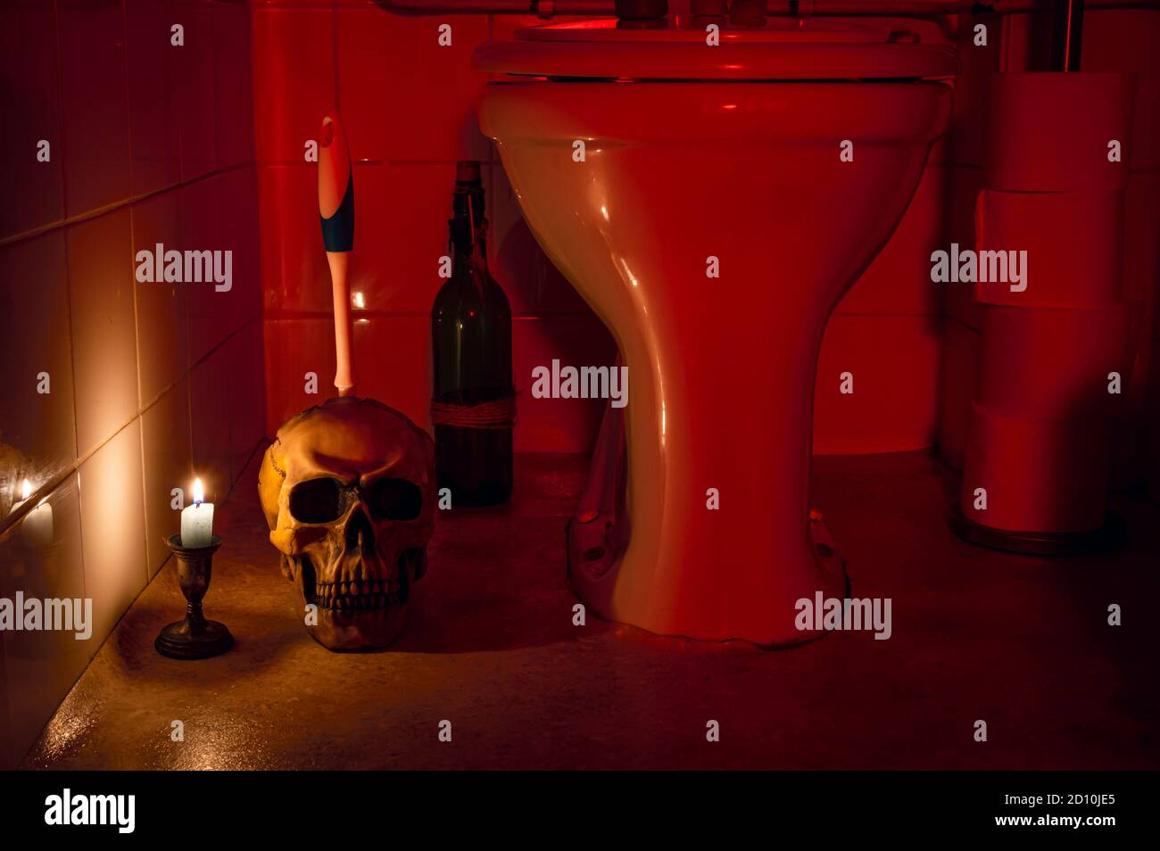Halloween restroom scene with a skull toilet brush holder and toilet. Scene is lit by candle light and daylight lamp with a red gel. Stock Photo
