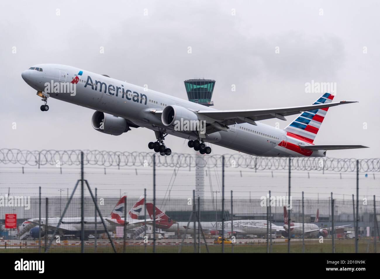 American Airlines Boeing 777 -300 jet airliner plane taking off in bad weather from London Heathrow Airport, UK. Security fence and air traffic tower Stock Photo