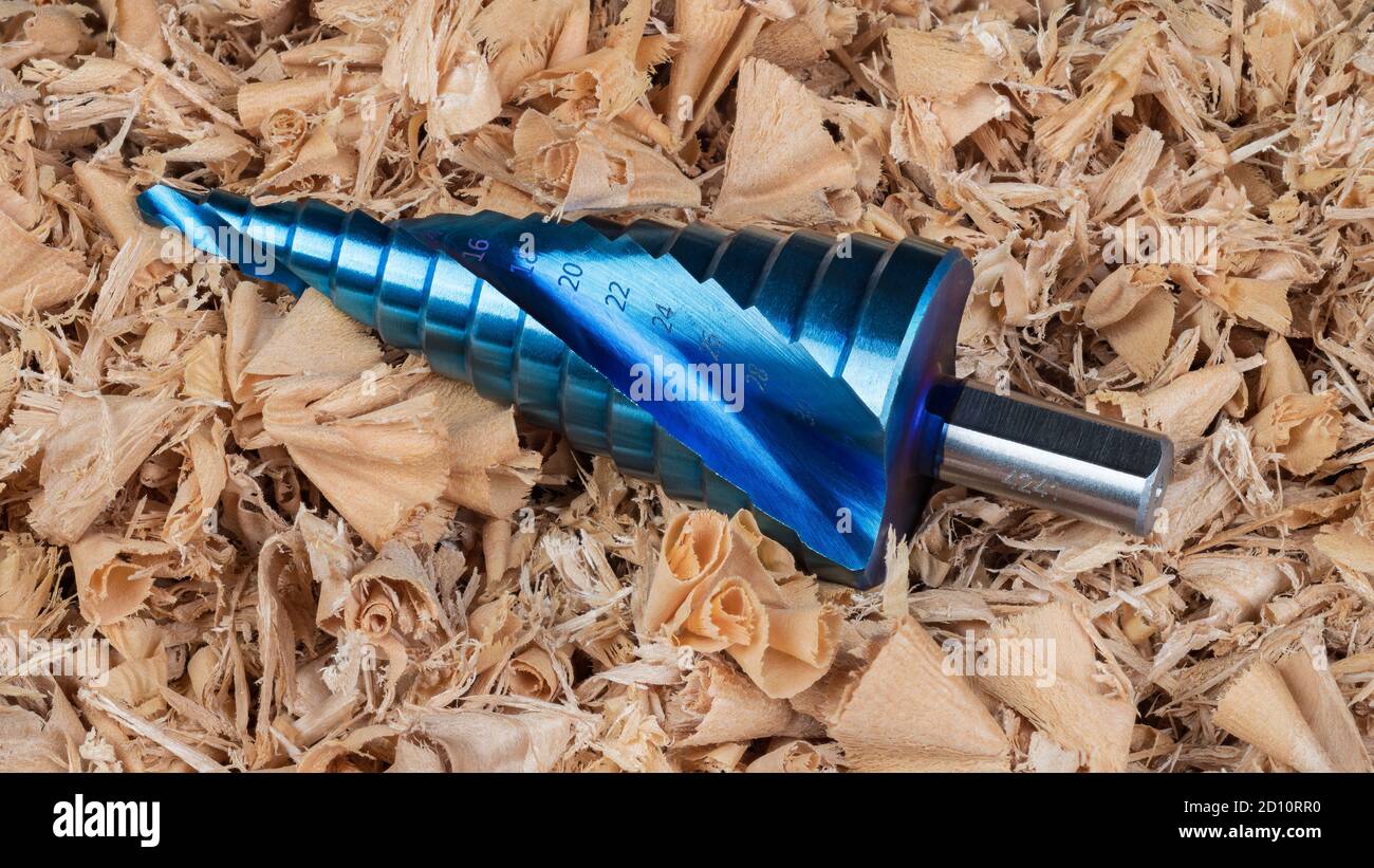 Cone step drill bit with blue nano coating on curled wood shavings pile. Sharp unibit from high quality steel. Woodworking chip machining cutting tool. Stock Photo
