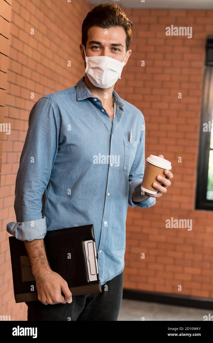 Portrait of middle east caucasian businessman with takeout coffee cup and clipboard in meeting room after lunch break. He wear face mask as new normal Stock Photo