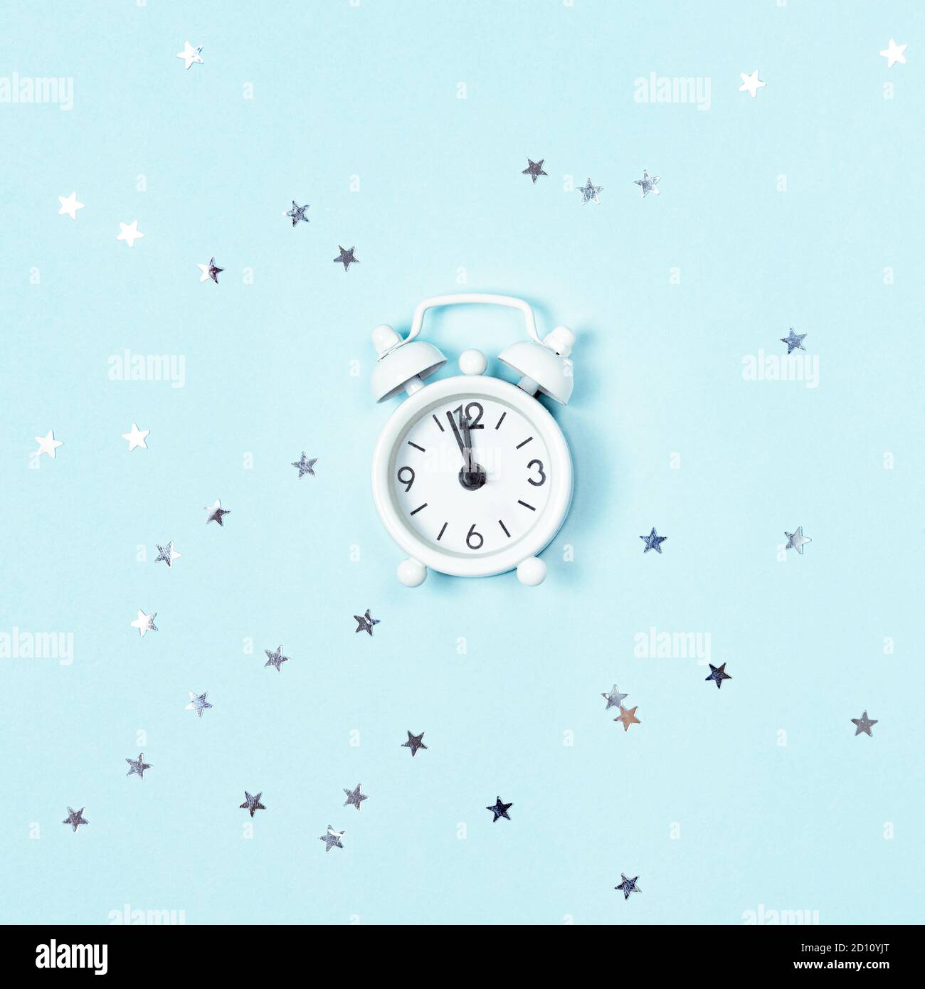 White vintage style alarm clock showing five minutes left untill midnight over blue background with silver stars sequins. New Year advent. Stock Photo
