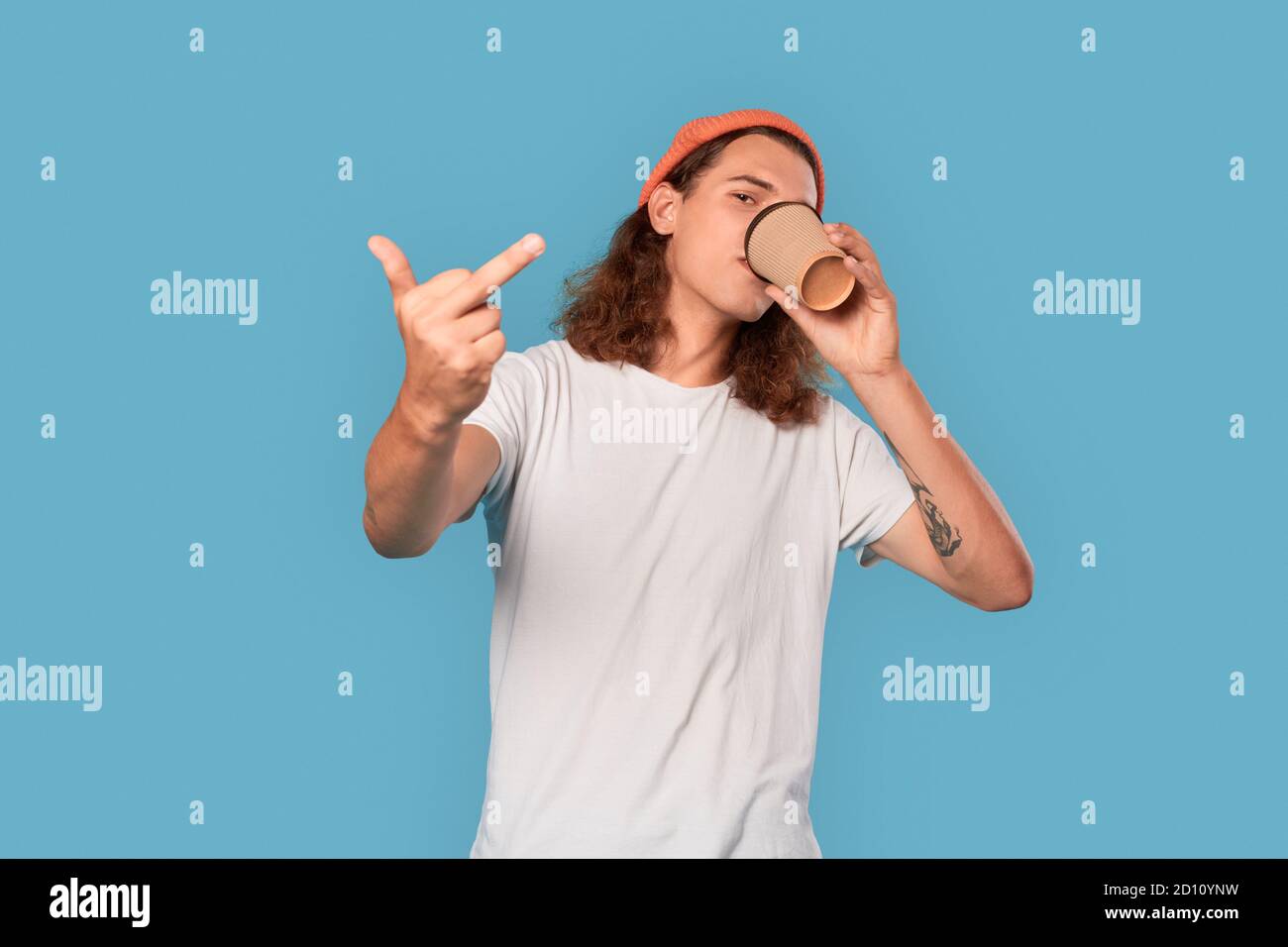 Freestyle. Curly-haired guy wearing hat standing isolated on blue drinking coffee showing middle finger cool Stock Photo