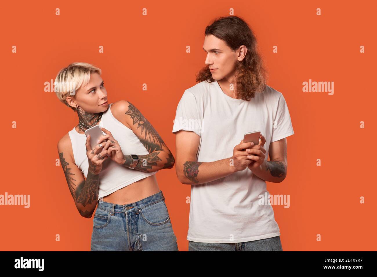Freestyle. Young tattooed couple standing isolated on orange with smartphones looking at each other nervous hiding information Stock Photo