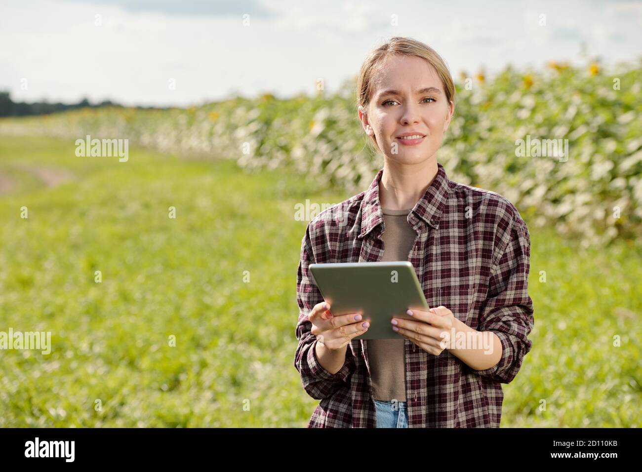 Young blond smiling female farmer in workwear using tablet against green field Stock Photo