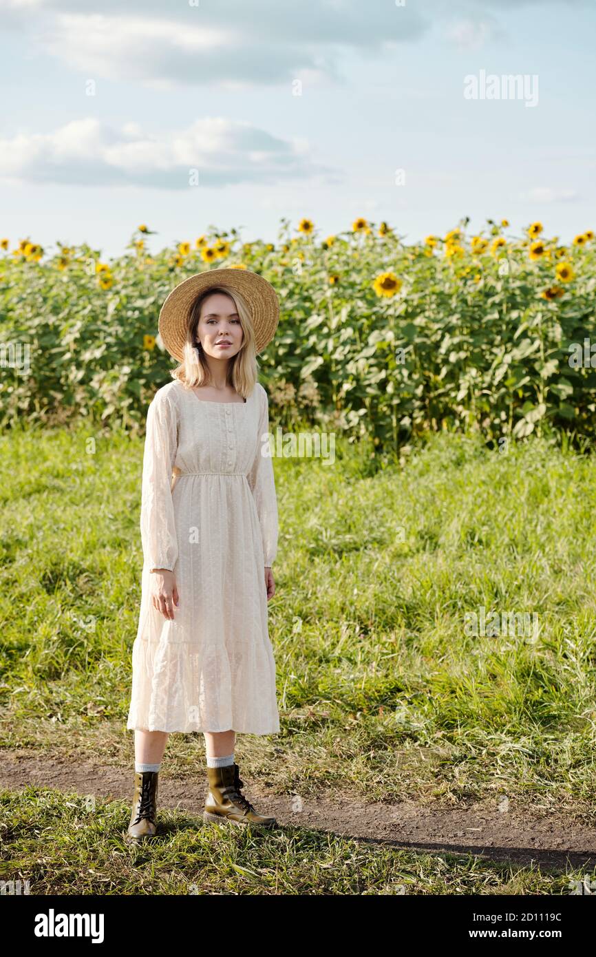 Happy young blond woman in hat and white romantic dress standing on green grass Stock Photo