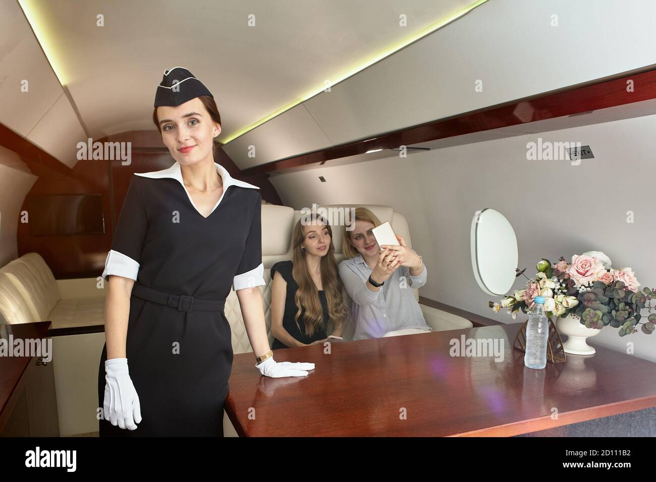Air hostess serves passengers inside the plane. Beautiful flight attendant in the passenger cabin of the aircraft on the background of young women in Stock Photo