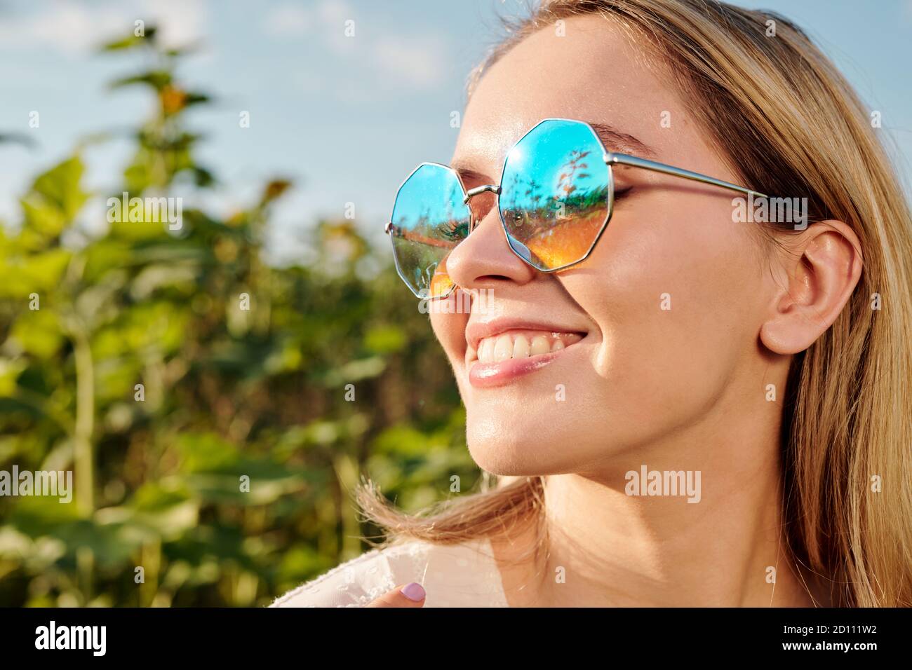 Face of smiling young blond woman in sunglasses standing against sunflowers Stock Photo