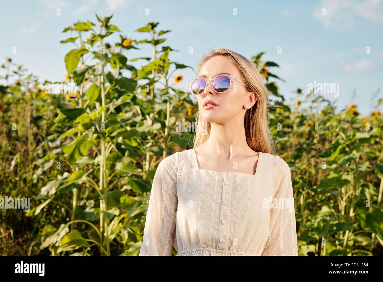 Blond young woman in sunglasses and white dress standing in front of camera Stock Photo