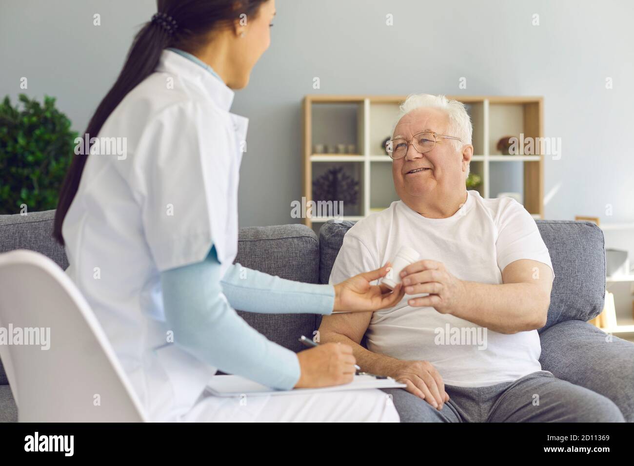 Young doctor visiting her senior patient at home, prescribing pills, supporting and cheering him up Stock Photo