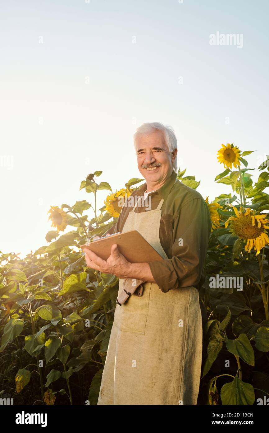 Senior laughing farmer in apron and shirt looking at you while reading document Stock Photo